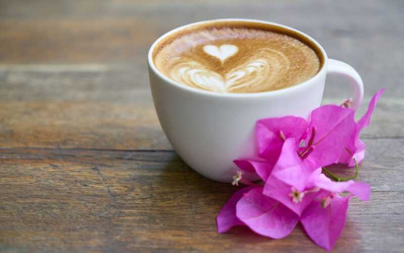 kopje koffie met in het koffieschuim een hartje gemaakt en naast het kopje ligt een roze bloem